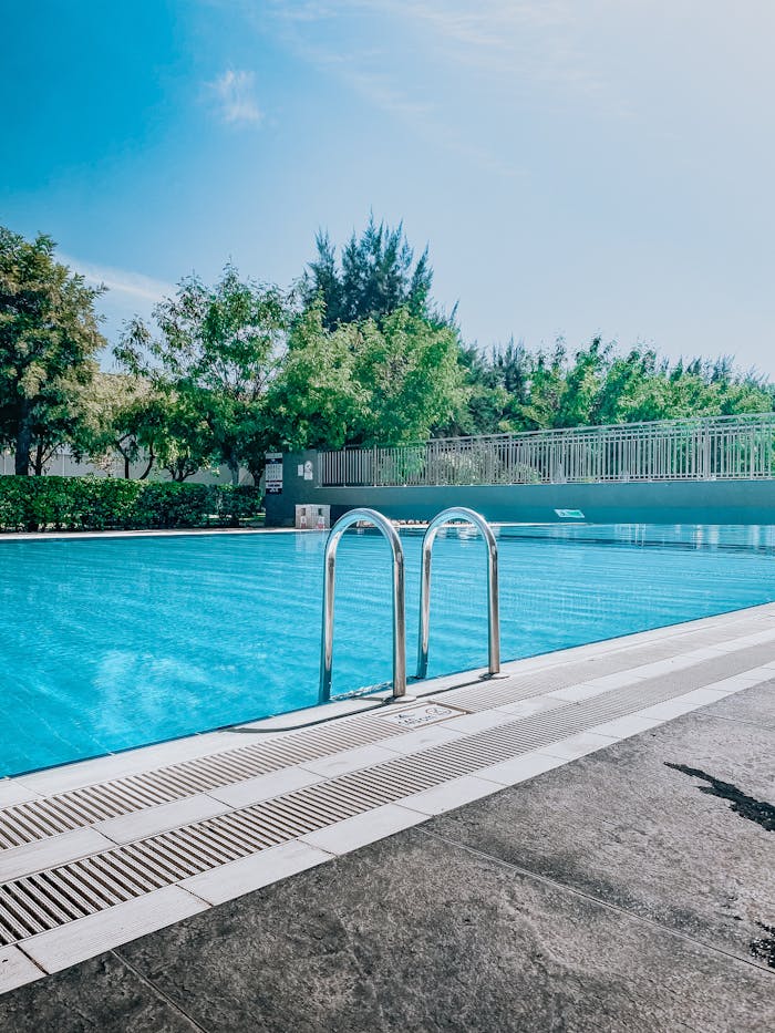 Home A serene outdoor swimming pool with clear blue water and lush greenery under a bright sky.